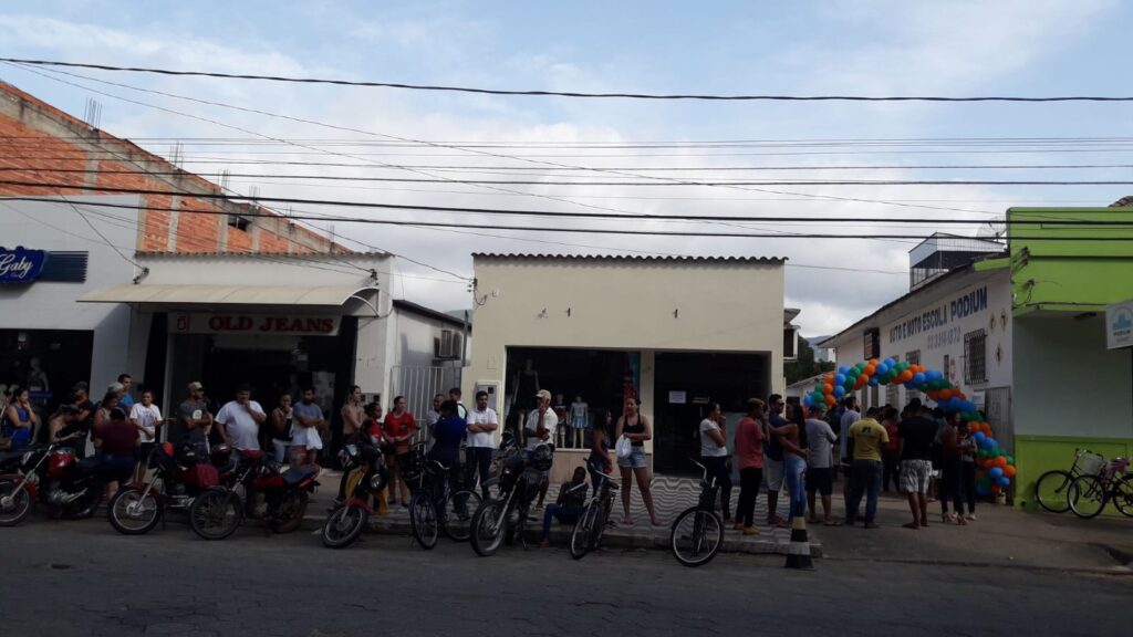Fila de interessados na calçada, com motos e bicicletas estacionadas e arco de balões na entrada da autoescola